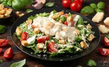 Fresh Caesar salad featuring grilled chicken breast, crispy croutons, romaine lettuce, tomatoes, and creamy dressing on a black plate.