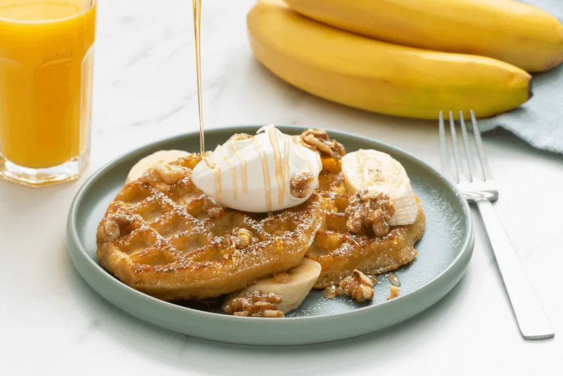 Plated French toast with caramelized exterior, topped with whipped cream and walnuts, served with fresh bananas and orange juice.