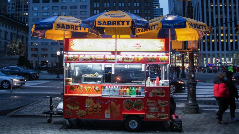 A red Sabrett food cart with yellow and blue umbrellas on an urban street corner, serving hot dogs and beverages to pedestrians.